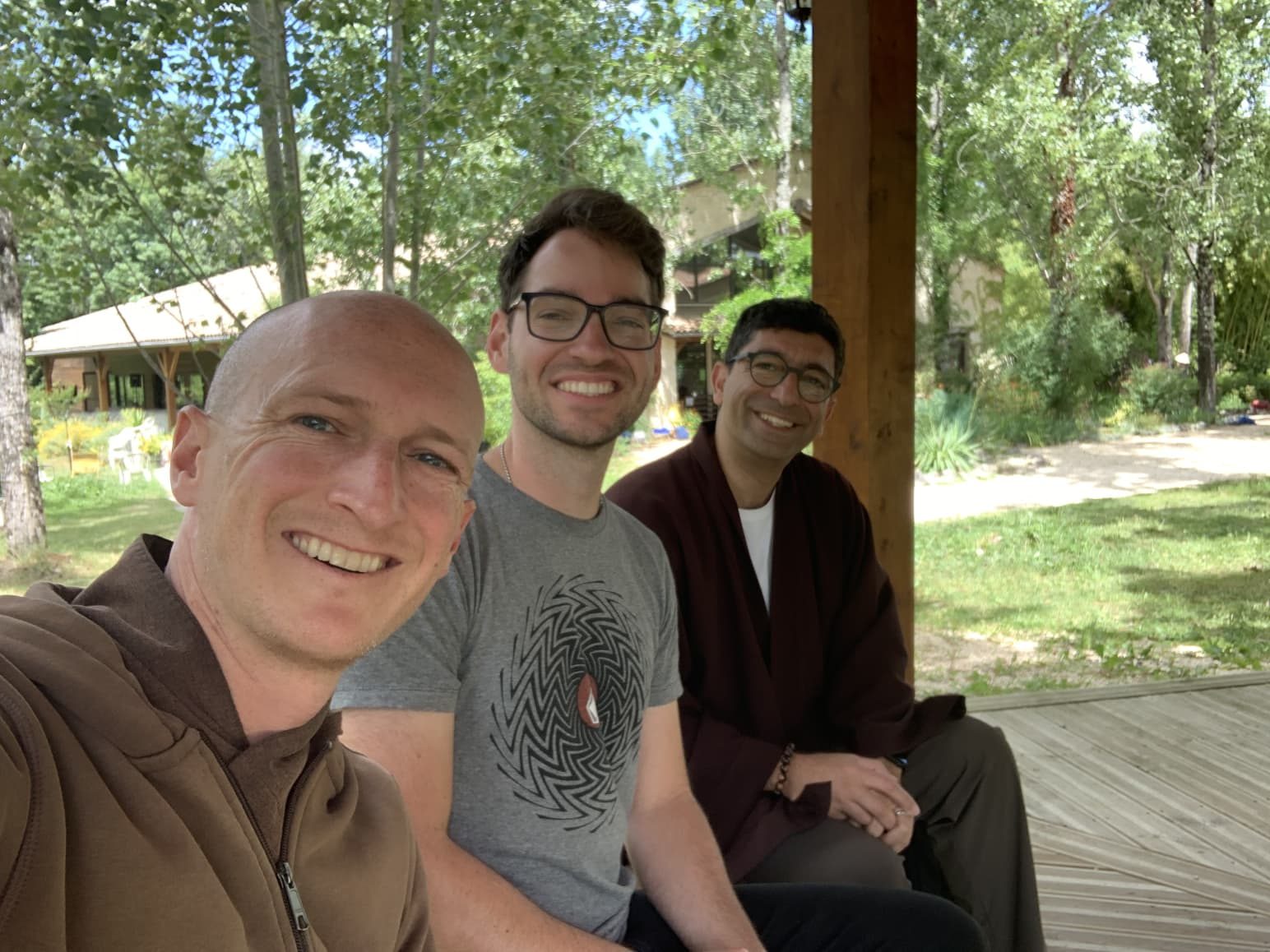 Three friends sitting together on a wooden veranda in the sunshine, smiling towards the camera, with trees and a low building behind them.
